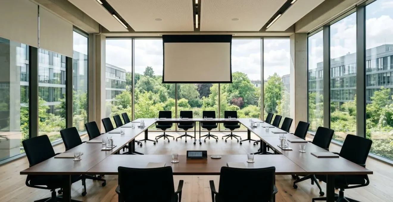 Salle de réunion contemporaine vide avec grandes fenêtres laissant entrer la lumière du jour, tables disposées en U, écran de projection et vue sur jardin extérieur